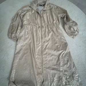 Beige regnjacka från Pureva - Säljer en beige regnjacka från Pureva. Den har ett glansigt material med ett subtilt mönster och dragkedja framtill. Jackan har trekvartsärmar och en hög krage för extra skydd mot regn. Perfekt för regniga dagar!