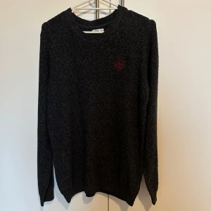 Mörkgrå stickad tröja från Gemo - Säljer en snygg och bekväm mörkgrå stickad tröja från Gemo. Den har ett coolt rött broderat märke på bröstet och är perfekt för kyligare dagar. Tröjan är långärmad och har en klassisk passform. Perfekt för både vardag och lite finare tillfällen!