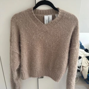 Beige fluffig tröja från Collusion - Supermysig beige tröja från Collusion! Den är fluffig och perfekt för kyliga dagar. Tröjan har en v-ringad design, vilket ger en trendig look. Perfekt för höst och vinter när du vill vara både varm och stilren.