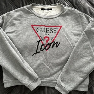 Grå sweatshirt från Guess - Snygg grå sweatshirt från Guess med deras ikoniska logga i rött och svart på framsidan. Tröjan har långa ärmar och en cropped passform. Använd några få gånger, därmed i mycket god skick.