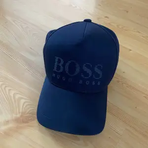 Säljer en snygg mörkblå keps från Hugo Boss. Den har en stilren design med 'BOSS' broderat på framsidan. Perfekt för både vardag och sportiga tillfällen. Justerbar passform för att sitta bekvämt hela dagen.