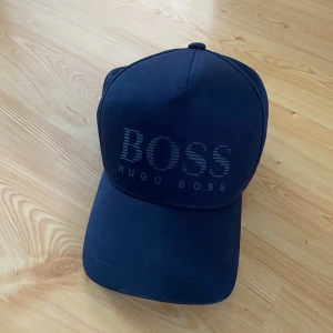 Mörkblå keps från Hugo Boss - Säljer en snygg mörkblå keps från Hugo Boss. Den har en stilren design med 'BOSS' broderat på framsidan. Perfekt för både vardag och sportiga tillfällen. Justerbar passform för att sitta bekvämt hela dagen.