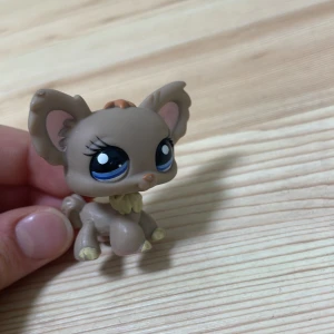 Brun figur från Littlest Pet Shop - Söt brun figur från Littlest Pet Shop med stora blå ögon och rosa detaljer i öronen. Figuren har en liten svans och en ljusare krage runt halsen. Perfekt för samlare eller som leksak för barn.