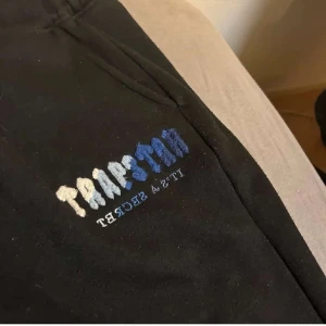 Svarta mjukisbyxor från trapstar - Säljer ett par svarta mjukisbyxor med broderad text i blått och vitt. Byxorna har snörning i midjan och dragkedjor vid fickorna. Perfekta för en avslappnad stil.