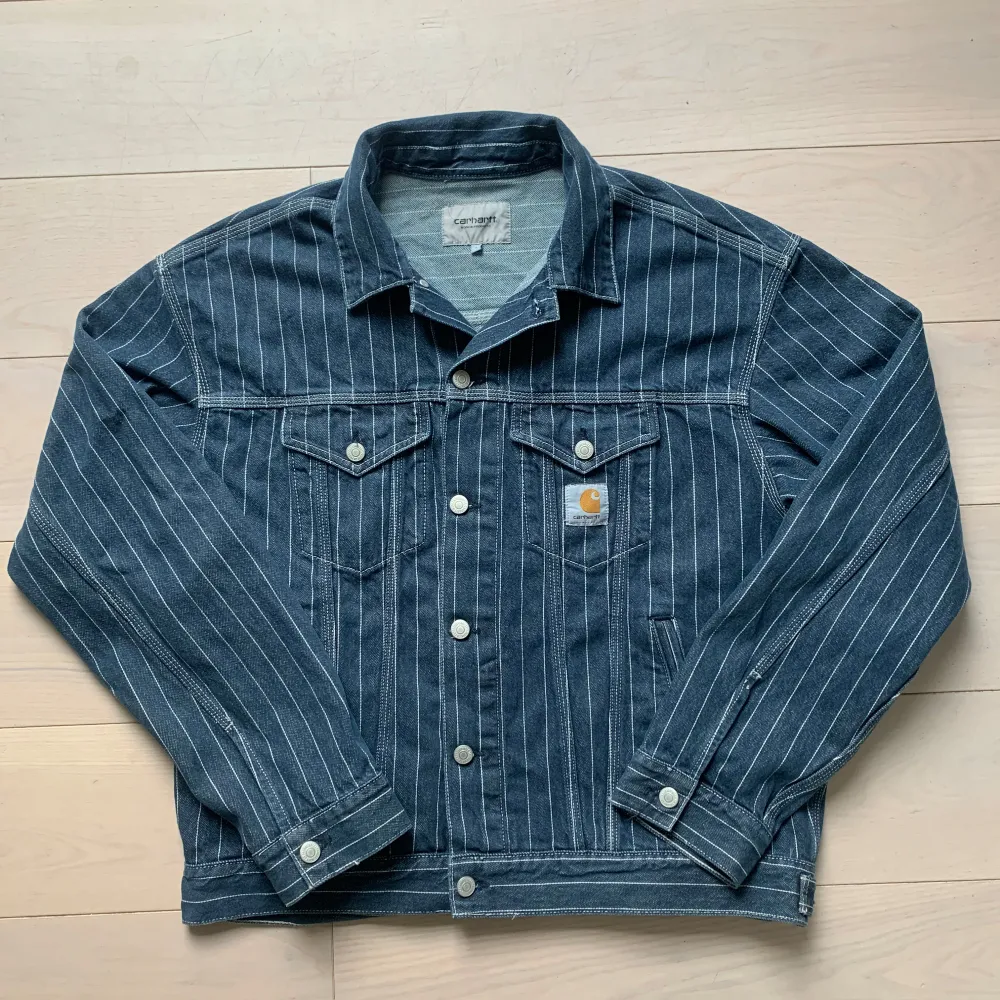 Säljer en snygg randig jeansjacka från Carhartt i blått med vita ränder. Jackan har en klassisk krage och knappar framtill samt på ärmsluten. Två bröstfickor med knappstängning och Carhartt-logga på vänster sida. Perfekt för en stilren look!. Takit.