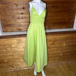Limegrön klänning med smala axelband - Snygg limegrön klänning med smala axelband och v-ringad design. Klänningen har en smockad midja som ger en fin passform och en luftig kjol som slutar vid anklarna. Perfekt för sommarens varma dagar!
