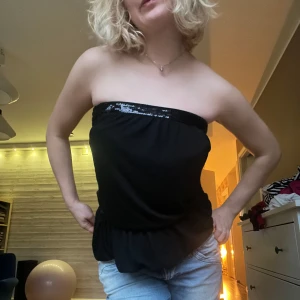 Svart tubtopp med paljettdetaljer - Snygg svart tubtopp med glittrande paljettdetaljer längs överkanten. Perfekt för en kväll ute eller en fest. Toppen har en elastisk passform som sitter bekvämt och stilrent.