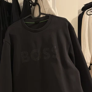 Svart tröja från BOSS - Säljer en stilren svart tröja från BOSS med texten 'BOSS' i glittriga detaljer på framsidan. Tröjan har en klassisk rund hals och långa ärmar. Perfekt för en avslappnad men ändå elegant look.