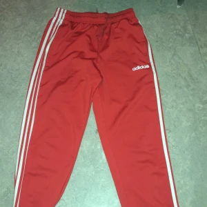 Röd träningsjacka från Adidas - Säljer en röd träningsjacka från Adidas med klassiska vita ränder längs ärmarna. Jackan har dragkedja framtill och två fickor. Perfekt för träning eller en sportig look.