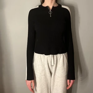 Svart och vit ribbad tröja med dragkedja som fungerar galant - Snygg svart ribbad tröja med vita detaljer längs ärmarna och en dragkedja framtill. Perfekt för en stilren look. Tröjan har en croppad passform och långa ärmar.