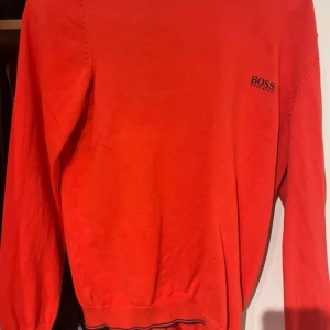 Röd tröja från Boss - Snygg röd tröja från Boss med broderad logga på bröstet. Tröjan har långa ärmar och ribbade muddar vid ärmslut och nederkant. Perfekt för en stilren look.