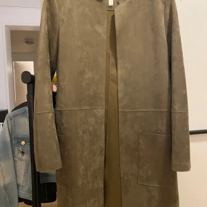 Olivgrön kappa från H&M - Snygg olivgrön kappa från H&M i ett stilrent och minimalistiskt snitt. Jackan har långa ärmar och stora fickor framtill, perfekt för en chic och modern look. 