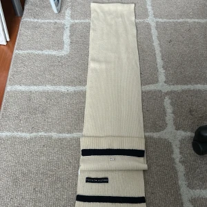 Beige halsduk från Tommy Hilfiger - Snygg beige halsduk från Tommy Hilfiger med subtilt märke i mitten. Den har svarta ränder vid ändarna som ger en stilren look. Perfekt för att hålla dig varm och trendig under kyliga dagar.