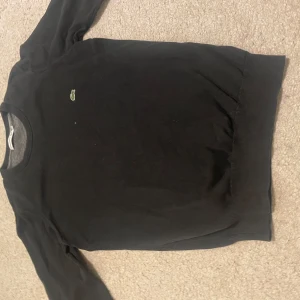 Svart tröja från Lacoste - Säljer en stilren svart tröja från Lacoste med det klassiska krokodilmärket på bröstet. Tröjan har långa ärmar och en rund halsringning. Perfekt för en avslappnad och snygg look.