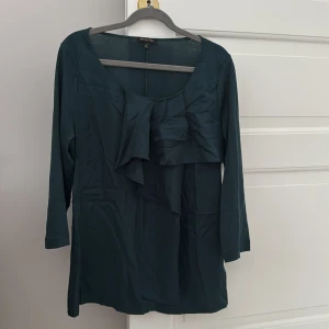 Mörkgrön blus från Massimo Dutti - Elegant mörkgrön blus från Massimo Dutti med volangdetaljer framtill. Blusen har långa ärmar och en rund halsringning 🌿