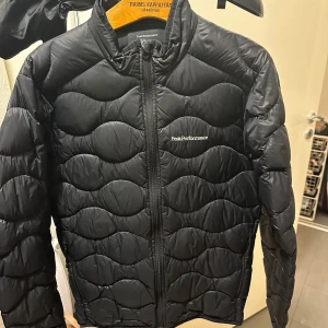 Svart dunjacka från Peak Performance - Säljer en svart dunjacka från Peak Performance med quiltat mönster. Jackan har en dragkedja framtill och är långärmad. Perfekt för kyligare dagar.
