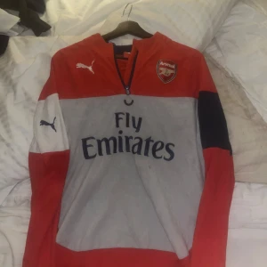 Röd och grå Arsenal-tröja från Puma - Säljer en snygg röd och grå Arsenal-tröja från Puma med klubbens logga och Fly Emirates-tryck. Tröjan har långa ärmar och en dragkedja vid halsen. Perfekt för fotbollsfans!