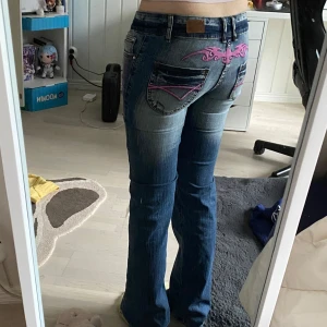 Blå jeans med rosa broderi från Crazy Age - Snygga blå jeans från Crazy Age med unikt rosa broderi på bakfickorna och framsidan. De har en bootcut-stil och är lågmidjade med dubbla knappar i midjan. Perfekta för en cool och avslappnad look. Helt nya och oanvända, lappen finns kvar!
