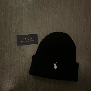 Svart mössa från Polo Ralph Lauren - Snygg svart mössa från Polo Ralph Lauren med broderad logotyp framtill. Perfekt för att hålla värmen under kyliga dagar. Klassisk design som passar till det mesta.