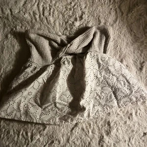Beige axelbandslös topp med spetsdetaljer - Säljer en söt beige axelbandslös topp med spetsdetaljer och broderade blommor. Toppen har en smockad överdel som ger en bra passform och en luftig nederdel. Perfekt för sommarens varma dagar!