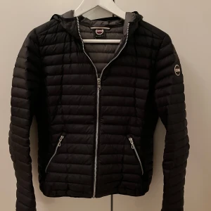 Svart dunjacka från Colmar - Snygg svart dunjacka från Colmar med dragkedja och huva. Jackan har en quiltad design och två praktiska fickor med dragkedjor. Perfekt för våren/ hösten och ger en stilren look. Inga defekter. Storlek 38, Colmar storlek 44