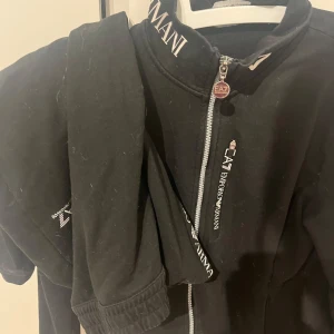 Svart jacka från Armani - Snygg svart jacka från Armani med dragkedja och logotypdetaljer. Jackan har långa ärmar och en hög krage med märkets namn. Perfekt för en stilren look.