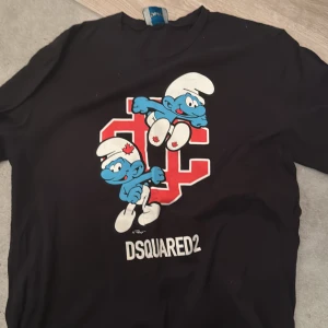 Svart Smurfarna t-shirt från Dsquared2 - Cool svart t-shirt från Dsquared2 med Smurfarna-tryck. Perfekt för dig som gillar unika och roliga designer. T-shirten har en stor röd bokstav och två smurfar på framsidan. Ett måste för alla Smurfarna-fans!