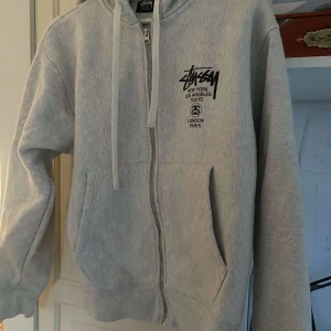 Grå hoodie från Stüssy - Säljer en grå hoodie från Stüssy med dragkedja och tryck på både fram- och baksidan. Hoodien har en justerbar huva med snören och en känguruficka. Perfekt för en avslappnad stil.