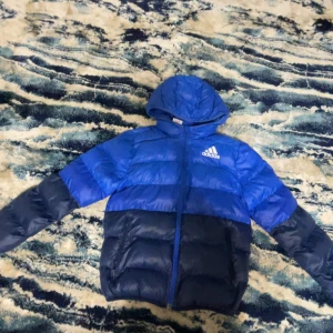 Blå barn jacka från Adidas - Säljer en blå pufferjacka från Adidas med huva och dragkedja. Jackan har en tvåtonad design i ljusblått och mörkblått. Perfekt för kyliga dagar.Passar bäst tiol barn 10-11år gammla (skriv för mer information)