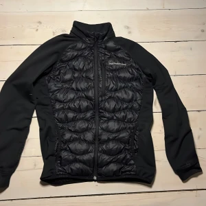 Svart Cardigen från Peak Performance - Säljer en svart Cardigen från Peak Performance med quiltade detaljer och dragkedja framtill. Jackan har långa ärmar och en stilren design, perfekt för kyligare dagar. Den är både funktionell och snygg, med en passform som sitter bra på kroppen.