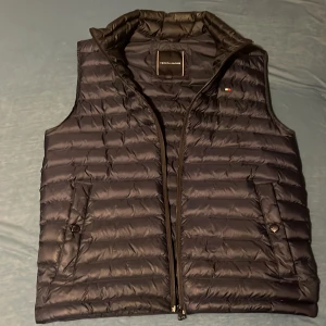Mörkblå navy dunväst från Tommy Hilfiger - Snygg svart dunväst från Tommy Hilfiger med dragkedja och två fickor framtill. Perfekt för lager-på-lager under kyliga dagar. Diskret logga på bröstet ger en stilren touch. 8.5av10 skick sparsamt använd köpt från Tommy hilfinger i Göteborg något år sedan för typ 2000kr 