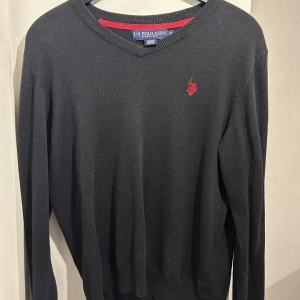 Svart tröja från US Polo Assn - Snygg svart tröja från US Polo Assn med röd broderad logga på bröstet. Tröjan har en v-ringad hals och långa ärmar, perfekt för en stilren look. Passar bra till både jeans och chinos. Tunn i materialet 