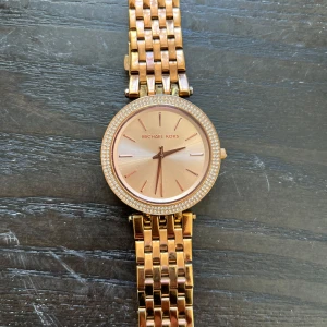 Roséguldfärgad klocka från Michael Kors - Elegant klocka i roséguld från Michael Kors med en glittrig ram runt urtavlan. Klockan har en länkarmband i metall och en stilren urtavla utan siffror. Perfekt för att ge en lyxig touch till din stil. Slitet skick i länk armbandet, se bilder, är ok skick runt uret, batteriet funkar, har ej några extra länkar, box ingår  :)   Skicka meddelande för mer info och bilder :)
