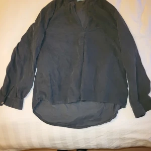 Grå långärmad manchesterskjorta - Säljer en stilren grå långärmad skjorta med en avslappnad passform. Skjortan har en enkel design utan synliga knappar framtill och är tillverkad i ett manchestertyg. Perfekt för en casual look.