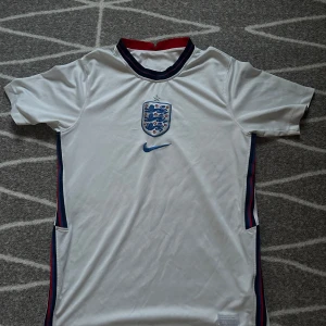 England fotbollströja - Säljer en vit fotbollströja från Nike med Englands landslagsemblem på bröstet. Tröjan har korta ärmar och en rund halsringning med mörkblå och röda detaljer. Perfekt för fotbollsfans som vill visa sitt stöd för laget!