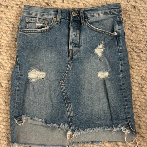 Jeanskjol från Lager  157 - Snygg jeanskjol från Lager  157 i en klassisk blå denimfärg. Kjolen har slitna detaljer och en rå kant nertill för en trendig look. Den har knappar framtill och praktiska fickor både fram och bak. Perfekt för en avslappnad stil.