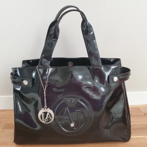 Svart glansig väska från Armani Jeans - Snygg svart glansig väska från Armani Jeans med dubbla handtag och ett stort AJ-logotyp på framsidan. Väskan har en avtagbar nyckelring med AJ-märke och knapplås på sidorna. Perfekt för att ge en stilren touch till din outfit. Ca 40×27, hand ca 36cm. Vanligt bruksslitage