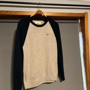 Grå och marinblå tröja från Hollister - Säljer en stilren grå och marinblå tröja från Hollister. Tröjan har långa ärmar och ett litet broderat logotyp på bröstet. Perfekt för en avslappnad stil.