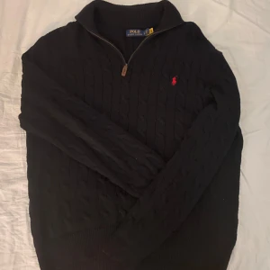 Svart stickad tröja från Ralph Lauren - Säljer en stilren svart stickad tröja från Ralph Lauren med dragkedja och det klassiska röda logotypen på bröstet. Tröjan har långa ärmar och en ribbad krage. Perfekt för kyligare dagar. 