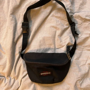 Svart midjeväska från Eastpak - Säljer en svart midjeväska från Eastpak med justerbart band och dragkedja. Perfekt för att hålla dina saker säkra och nära till hands. Väskan har en enkel och stilren design med Eastpak-loggan på framsidan.