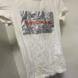 Vit t-shirt från Calvin Klein Jeans - Säljer en vit t-shirt från Calvin Klein Jeans med ett stort silverfärgat tryck och röd text på bröstet. T-shirten har korta ärmar och en klassisk rund halsringning. Perfekt för en stilren och modern look.