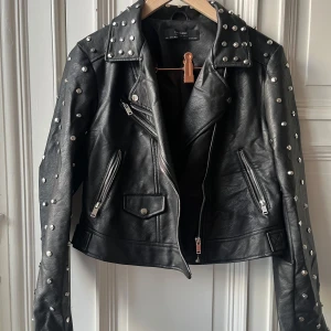 Svart skinnjacka med nitar från Zara - Snygg svart fejk skinnjacka från Zara med coola nitar på axlar och ärmar. Jackan har en unik broderad dödskalle på ryggen och dragkedjor på fickorna. Perfekt för att ge din outfit en edgy touch. Använd få tal gånger, inköpt för 6 år sedan, har blivit skrynklig efter ha varit i förrådet, liten i storlek och kort   Hund finns i hemmet, Skicka meddelande för mer info och bilder :)