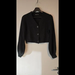 Svart croppad ribbad kofta - Svart kofta från med ribbad struktur och knappar framtill. Croppad. Hund finns i hemmet.