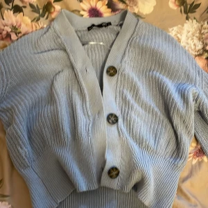 Ljusblå kofta från Lager 157 - Säljer en ljusblå kofta från Lager 157 i modellen Giselle. Koftan är stickad med stora knappar framtill och har långa ärmar. Perfekt för en stilren och bekväm look. Tillverkad i en blandning av bomull och akryl.