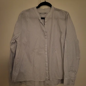 Randig henley-skjorta - Säljer en stilren randig henley-skjorta i ljusblått och vitt. Skjortan har långa ärmar och en klassisk krage utan knappar. Perfekt för en avslappnad men ändå snygg look.