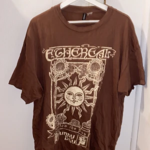 Brun t-shirt med soltryck från H&M Divided - Säljer en brun t-shirt från H&M Divided med ett coolt tryck av en sol och texten 'Follow the Sun, Sunny Days'. Perfekt för en avslappnad stil. T-shirten har korta ärmar och en rund halsringning.