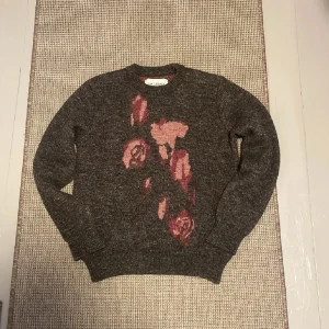 Our Legacy Tröja - Säljer en snygg grå stickad tröja från Our Legacy med ett unikt blommönster i rosa nyanser på framsidan. Tröjan är tillverkad i en ull- och linneblandning, vilket gör den både varm och bekväm. Perfekt för kyligare dagar! Nypris ligger på ungefär 4 tusen! Hör gärna av er vid flera frågor eller funderingar! 