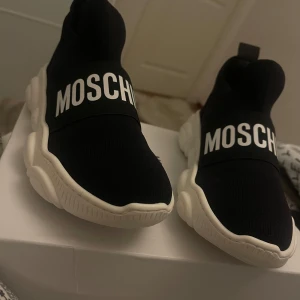 Svarta sneakers från Moschino - Snygga svarta sneakers från Moschino med vit sula och stort Moschino-tryck över ovansidan. Skorna har en unik sula med björnformad design undertill. Perfekta för en trendig look. Säljer skorna pågrund av fel storlek.