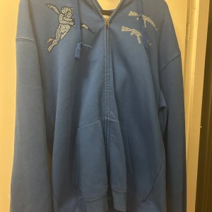 Blå hoodie från One of One - Säljer en helt oanvänd limiterad blå hoodie från One of One med dragkedja och unikt mönster av en figur med vingar och gevär på bröstet. Tröjan har långa ärmar och en bekväm passform. Perfekt för en avslappnad stil.