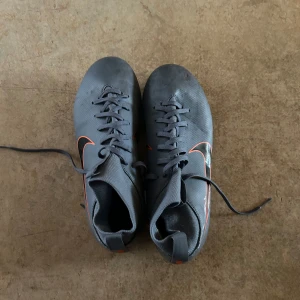 Grå Nike Mercurial fotbollsskor - Säljer ett par grå Nike Mercurial fotbollsskor med orange detaljer och snörning. Skorna har en strömlinjeformad design med dobbar för optimalt grepp på planen. Perfekta för fotbollsspelare som vill ha stil och funktionalitet.